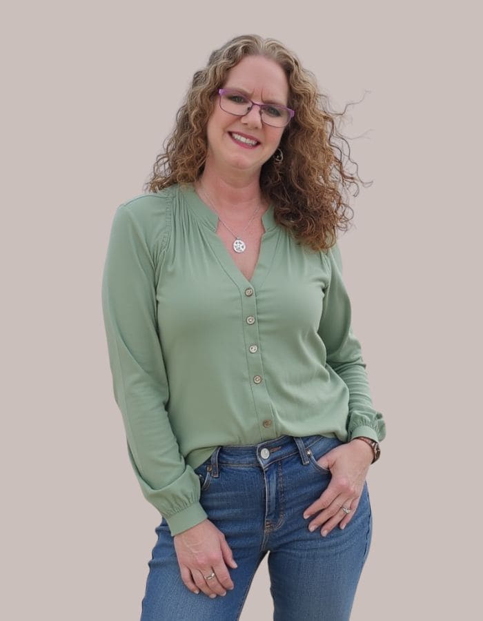 Woman with curly brown hair and glasses smiling at the camera while standing against a light neutral background. She is wearing a sage green button-front blouse, blue jeans, a silver necklace, and a watch, with one hand resting in her pocket and the other at her side.